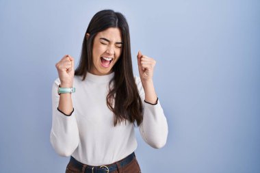 Young brunette woman standing over blue background excited for success with arms raised and eyes closed celebrating victory smiling. winner concept. 