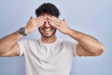 Hispanic man with beard standing over white background covering eyes with hands smiling cheerful and funny. blind concept. 