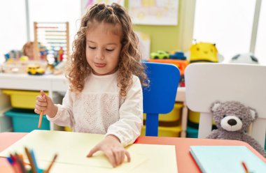 Sevimli sarışın anaokulu öğrencisi anaokulunda masaya oturmuş kağıda resim çiziyor.