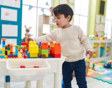 Adorable hispanic boy playing with construction blocks standing at kindergarten
