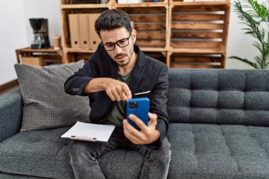Young hispanic psychologist man doing therapy on video call with smartphone pointing with finger to the camera and to you, confident gesture looking serious 