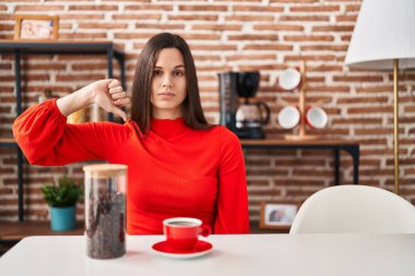 Young hispanic woman drinking coffee at home with angry face, negative sign showing dislike with thumbs down, rejection concept 