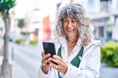 Sokakta akıllı telefon kullanan orta yaşlı bir kadın gülümsüyor.