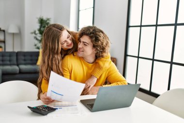 Genç beyaz çift, evde dizüstü bilgisayar kullanarak aile muhasebesi yaparak birbirlerine sarılıyorlar.