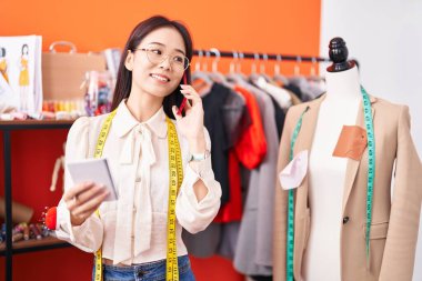 Young chinese woman tailor talking on smartphone reading notebook at atelier