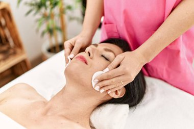 Middle age hispanic woman having facial treatment cleaning face at beauty center