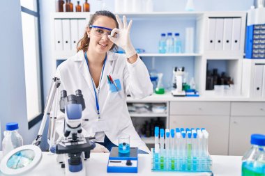 Young hispanic woman working at scientist laboratory doing ok gesture with hand smiling, eye looking through fingers with happy face. 