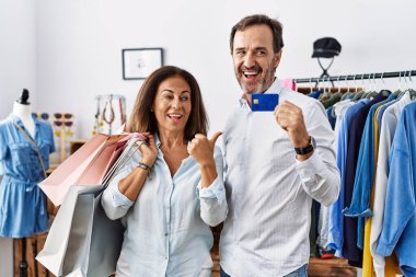 Hispanic middle age couple holding shopping bags and credit card smiling with happy face looking and pointing to the side with thumb up. 