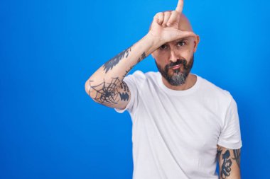 Hispanic man with tattoos standing over blue background making fun of people with fingers on forehead doing loser gesture mocking and insulting. 