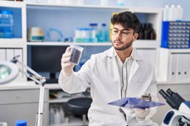 Young hispanic man scientist weighing sample reading document at laboratory