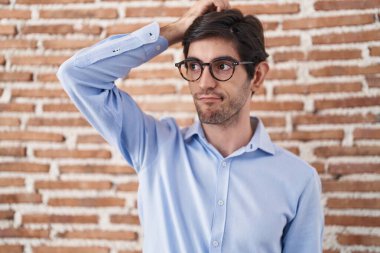 Young hispanic man standing over brick wall background confuse and wondering about question. uncertain with doubt, thinking with hand on head. pensive concept. 