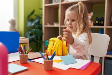 Anaokulunda elinde kalem tutarak gülümseyen sevimli sarışın kız anaokulu öğrencisi.