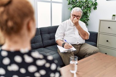 Senior psychologist man at consultation office smelling something stinky and disgusting, intolerable smell, holding breath with fingers on nose. bad smell 