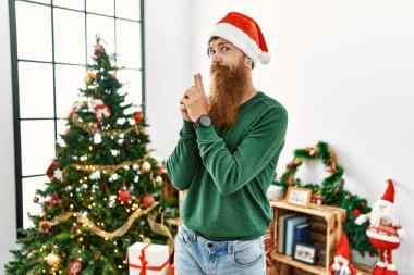 Redhead man with long beard wearing christmas hat by christmas tree holding symbolic gun with hand gesture, playing killing shooting weapons, angry face 