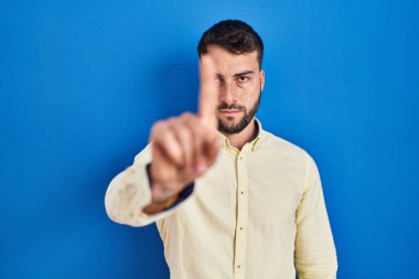 Handsome hispanic man standing over blue background pointing with finger up and angry expression, showing no gesture 