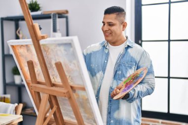 Sanat stüdyosunda kendine güvenen genç Latin sanatçı gülümsüyor.