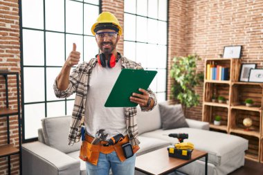 Evde çalışan sakallı, mutlu ve olumlu gülümseyen, başparmağı mükemmel ve onay işareti yapan genç İspanyol bir adam. 