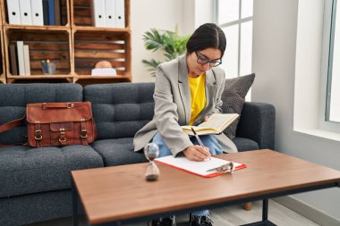 Young hispanic woman psychologist writing on clipboard reading book at clinic