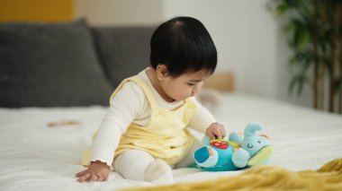Adorable hispanic baby playing with elephant doll sitting on bed at bedroom