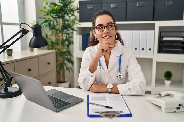 医師の制服を着た若いヒスパニック系の女性と顎の質問について考える上で手で聴診器 集中的な表現 笑顔で思いやりのある顔 疑わしい概念 — ストック写真