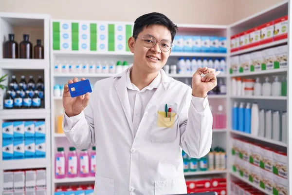 Young asian man working at pharmacy drugstore holding credit card smiling happy pointing with hand and finger to the side