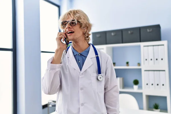 Young blond man doctor smiling confident talking on smartphone at clinic