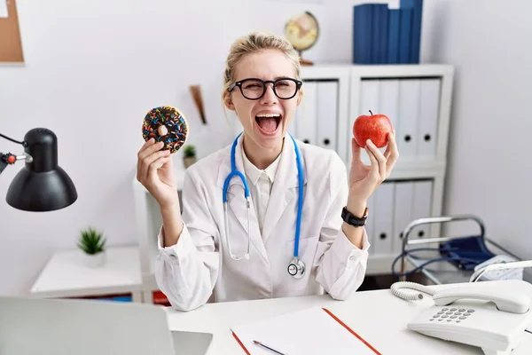 Young Doctor Woman Holding Red Apple Donut Clinic Celebrating Crazy —  Fotos de Stock
