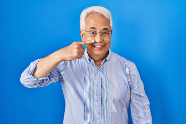 Hispanic senior man wearing glasses pointing with hand finger to face and nose, smiling cheerful. beauty concept