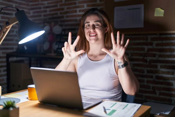 Brunette woman working at the office at night showing and pointing up with fingers number eight while smiling confident and happy.