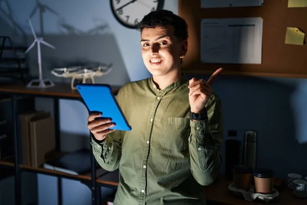 Non binary person using touchpad device at night with a big smile on face, pointing with hand finger to the side looking at the camera.