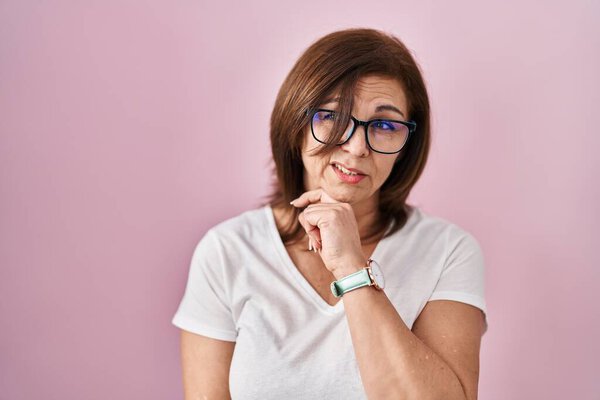 Middle age hispanic woman standing over pink background thinking worried about a question, concerned and nervous with hand on chin 