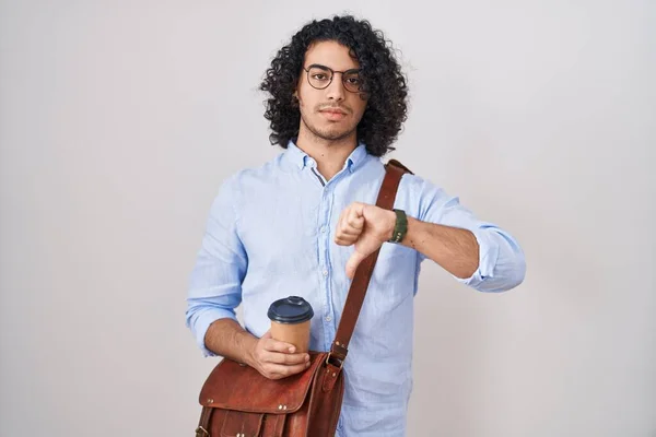 Hispanic man with curly hair drinking a cup of take away coffee looking unhappy and angry showing rejection and negative with thumbs down gesture. bad expression.