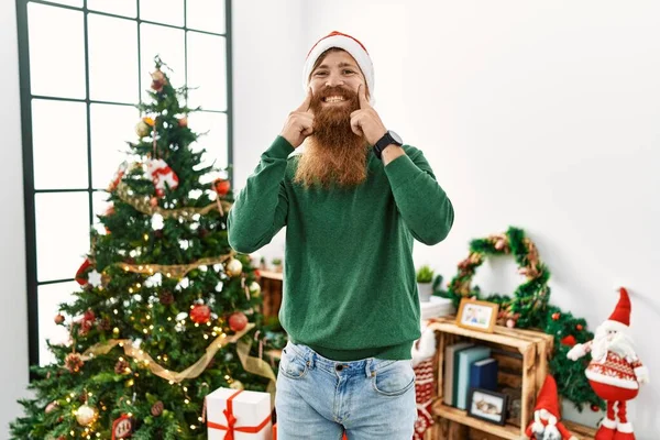 長いです髭を生やした男を身に着けていますクリスマスの帽子によってクリスマスツリー笑顔でオープン口 指指すと明るい笑顔を強制 — ストック写真