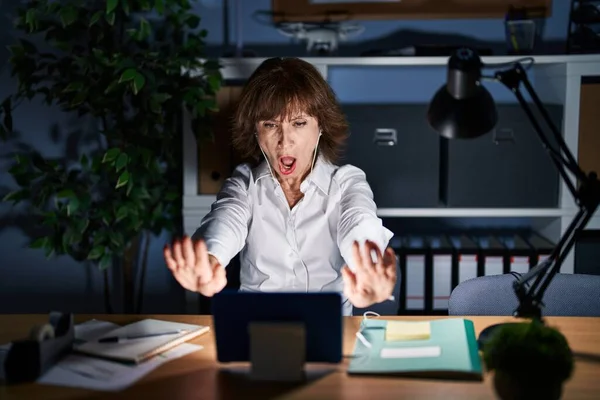Middle age woman working at the office at night doing stop gesture with hands palms, angry and frustration expression