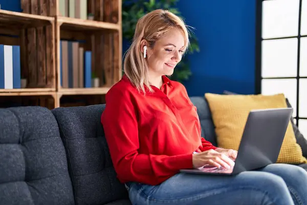 年轻的金发女人坐在家里的桌子上 手里拿着笔记本电脑和耳机 — 图库照片