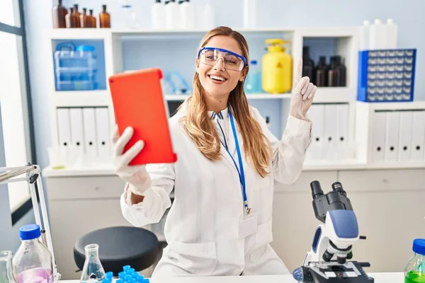 Young blonde scientist woman on video call working at laboratory smiling happy pointing with hand and finger to the side