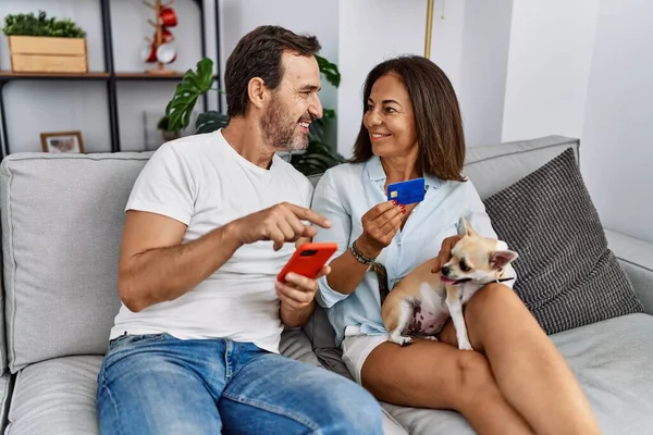 中年男性と女性カップル使用スマートフォンとクレジットカード座って上のソファに犬とともに自宅 — ストック写真