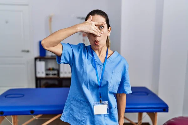 Mladá Hispánka Uniformě Fyzioterapeutky Stojící Klinice Vykukující Šoku Zakrývající Obličej — Stock fotografie