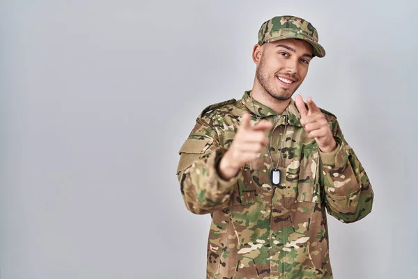 Young hispanic man wearing camouflage army uniform pointing fingers to camera with happy and funny face. good energy and vibes.