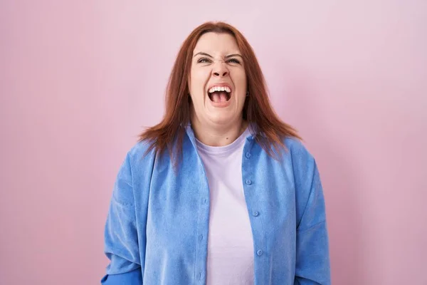 Young hispanic woman with red hair standing over pink background angry and mad screaming frustrated and furious, shouting with anger. rage and aggressive concept.