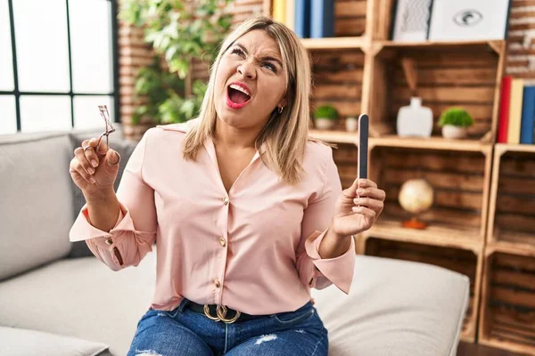 Young hispanic woman holding eye lashes curler and nail file angry and mad screaming frustrated and furious, shouting with anger looking up.