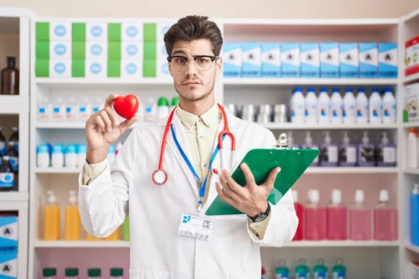 Young hispanic man working at pharmacy drugstore holding heart skeptic and nervous, frowning upset because of problem. negative person.
