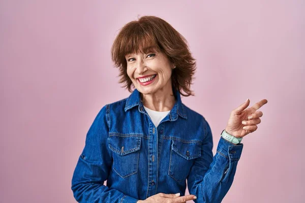 Middle age woman standing over pink background with a big smile on face, pointing with hand finger to the side looking at the camera.