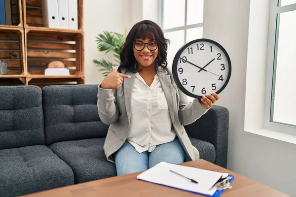 Hispanic woman working at therapy office holding clock pointing finger to one self smiling happy and proud