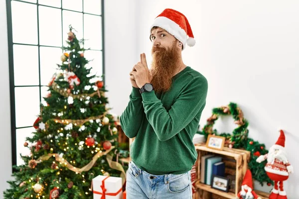 Redhead man with long beard wearing christmas hat by christmas tree holding symbolic gun with hand gesture, playing killing shooting weapons, angry face