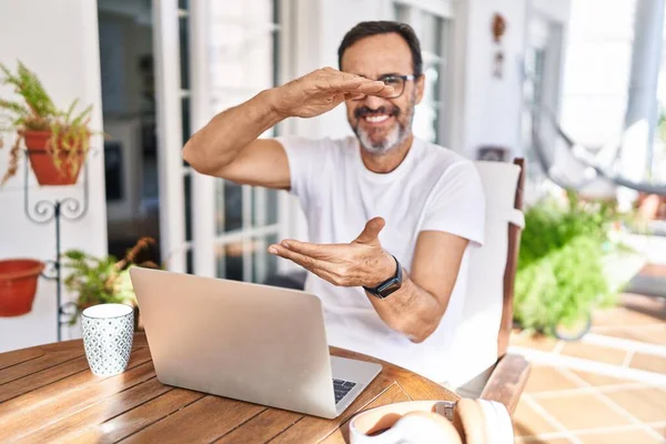 Middle age man using computer laptop at home gesturing with hands showing big and large size sign, measure symbol. smiling looking at the camera. measuring concept.