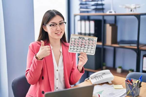 Chinese young woman working at the office holding holidays calendar smiling happy and positive, thumb up doing excellent and approval sign