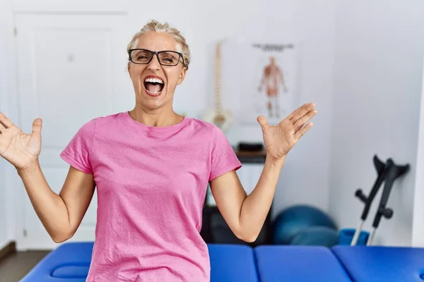 Middle age blonde woman at pain recovery clinic crazy and mad shouting and yelling with aggressive expression and arms raised. frustration concept.