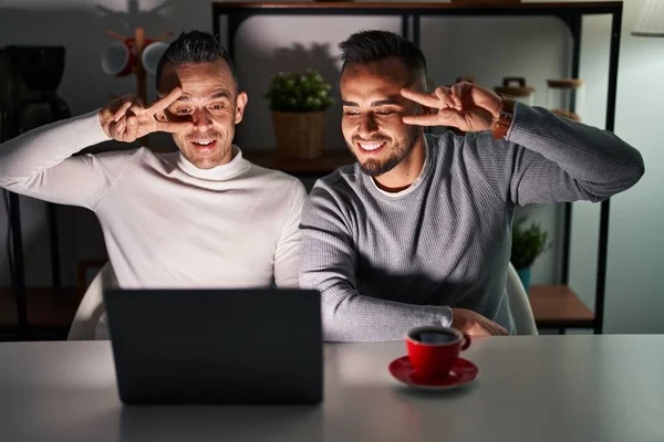 Homosexual couple using computer laptop doing peace symbol with fingers over face, smiling cheerful showing victory