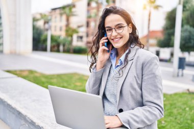 Genç İspanyol kadın yönetici dizüstü bilgisayar kullanıyor ve caddede akıllı telefondan konuşuyor.